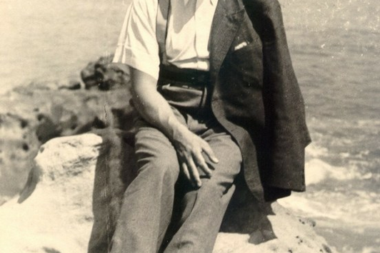 Ein Foto eines Herrn, während er auf einem Stein sitzt, hinter ihm befindet sich Wasser, See oder Meer.