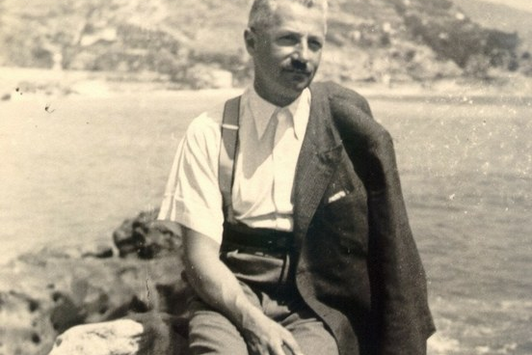 Ein Foto eines Herrn, während er auf einem Stein sitzt, hinter ihm befindet sich Wasser, See oder Meer.