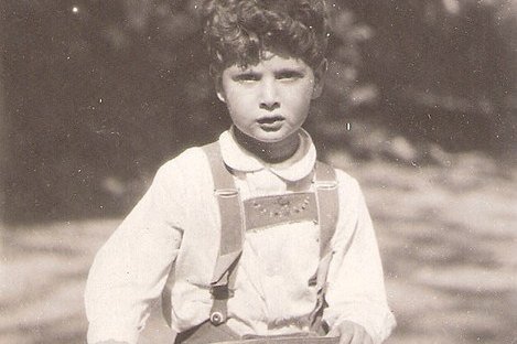 Ein junger Bub in Lederhose steht auf einem Roller aus Holz