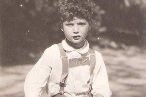 Ein junger Bub in Lederhose steht auf einem Roller aus Holz