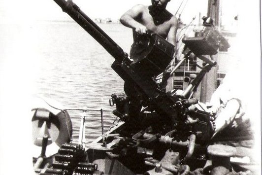 Ein junger Mann auf einem Kriegsschiff hält die Munitionstrommel einer Kanone