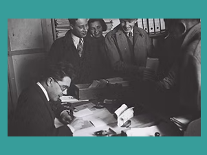 Ein Herr schreibt auf einem Schreibtisch, er ist umgeben von anderen Menschen