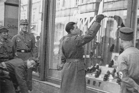 Uniformierte Nationalsozialisten stehen vor einem Schaufenster eines Geschäfts, einer von ihnen beschmiert dieses mit der Aufschrift Jude