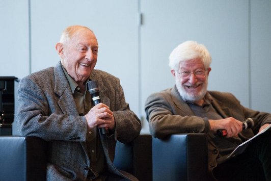 Zwei ältere Herren sitzen nebeneinander und halten Mikrofone in ihren Händen