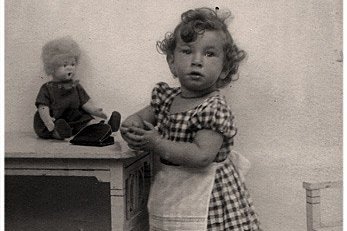 Ein kleines Mädchen lehnt sich an einen Tisch, auf dem Tisch befindet sich eine Spielzeugpuppe