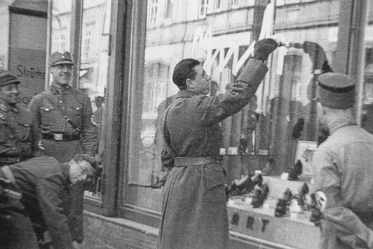 Uniformierte Nationalsozialisten stehen vor dem Schaufenster eines Geschäfts, einer von ihnen beschmiert dieses mit der Aufschrift Jude