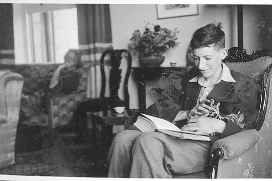 Ein junger Mann sitzt in einem Sessel und hat ein Buch auf dem Schoß. Er hält eine Katze in den Armen