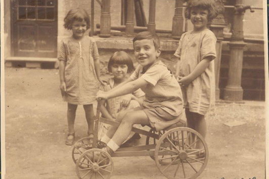 Vier Kinder spielen in einem Innenhof, ein junger Bursche sitzt auf einem Holzwagen
