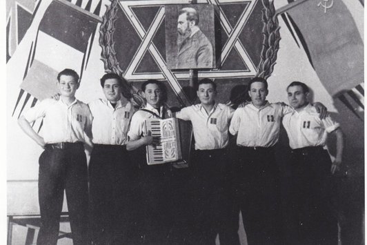 eine Gruppe junger Männer, einer hält ein Akkordeon, über ihnen befindet sichder Davidstern mit einem Portrait Theodor Herzels