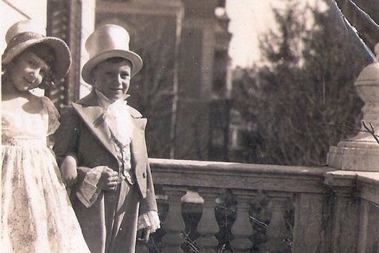 Ein junges Mädchen und ein junger Bub, formal gekleidet auf einem Balkon