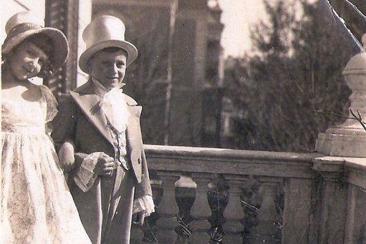 Ein junges Mädchen und ein junger Bub, formal gekleidet auf einem Balkon